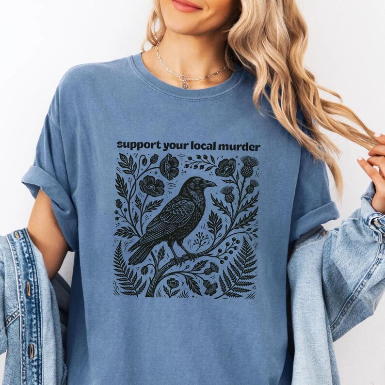 May include: A blue t-shirt featuring a black graphic print of a crow perched on a branch surrounded by flowers and leaves. The text "support your local murder" is printed in black above the illustration. The shirt has short sleeves and a crew neck.