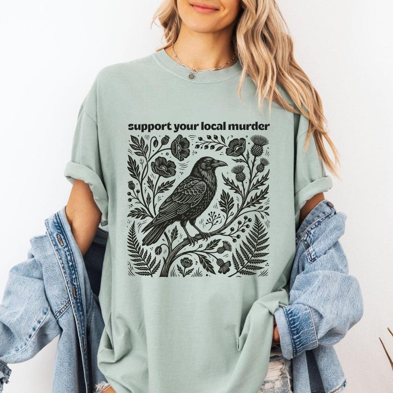 May include: A sage green t-shirt featuring a black graphic print of a crow perched on a branch surrounded by botanical elements like flowers and ferns. The text "support your local murder" is printed in black above the crow illustration.