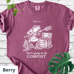 May include: Berry-colored t-shirt with a humorous graphic of a frog holding a pitchfork, standing near a wooden chest. Text on the shirt reads "That's it." and "You're going in the COMPOST."