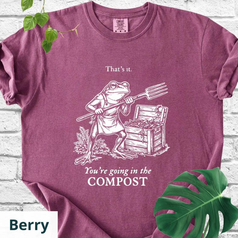 May include: Berry-colored t-shirt with a humorous graphic of a frog holding a pitchfork, standing near a wooden chest. Text on the shirt reads "That's it." and "You're going in the COMPOST."