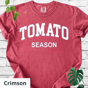 K&ouml;nnte beinhalten: Karmesinrotes T-Shirt mit dem wei&szlig;en Schriftzug "TOMATO SEASON". Das Shirt hat einen Rundhalsausschnitt und kurze &Auml;rmel. Das Kleidungsst&uuml;ck ist einfarbig und hat ein leicht verwaschenes Aussehen.
