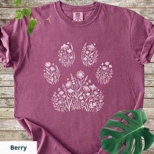 May include: A berry-colored short-sleeve t-shirt featuring a white graphic print of a paw print shape composed of various wildflowers and small butterflies. The shirt has a crew neckline and a label at the inner collar reading Comfort Colors.