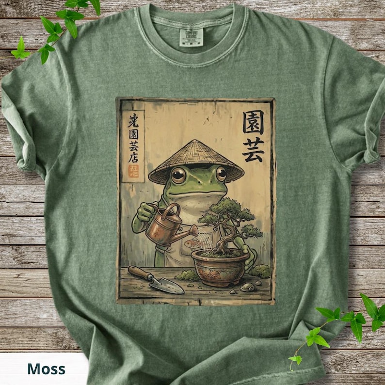 May include: Moss green t-shirt featuring a frog in a conical hat watering a bonsai tree. The frog illustration is set against a wooden background with Japanese text. A small trowel is also visible.