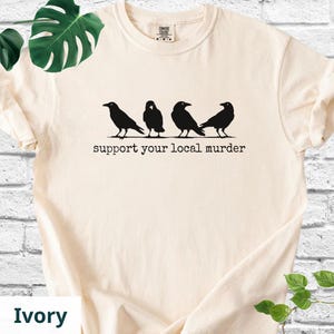 May include: An ivory short-sleeved t-shirt featuring a black graphic of four crows standing in a row. Below the birds, the text reads "support your local murder" in a typewriter-style font. The shirt has a crew neckline and a standard fit.