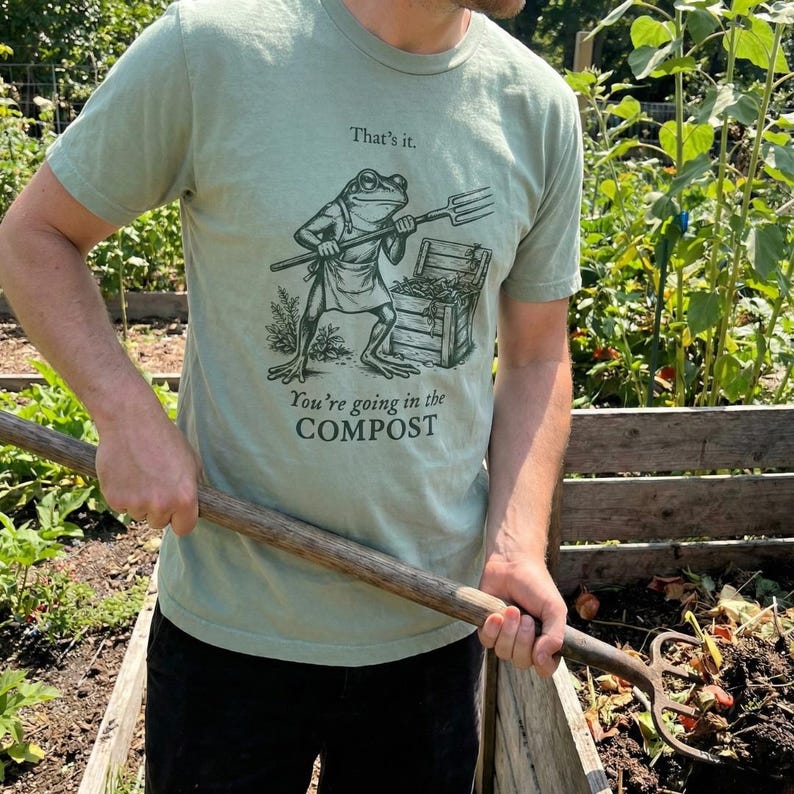 May include: Sage green t-shirt featuring a frog illustration with a pitchfork and apron, standing near a compost bin. The text on the shirt reads "That's it." and "You're going in the COMPOST."