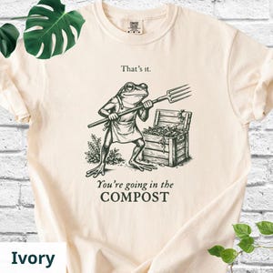 May include: Ivory-colored t-shirt featuring a humorous illustration of a frog holding a pitchfork, standing near a wooden compost bin. The text "That's it." and "You're going in the COMPOST" are printed above and below the frog graphic.