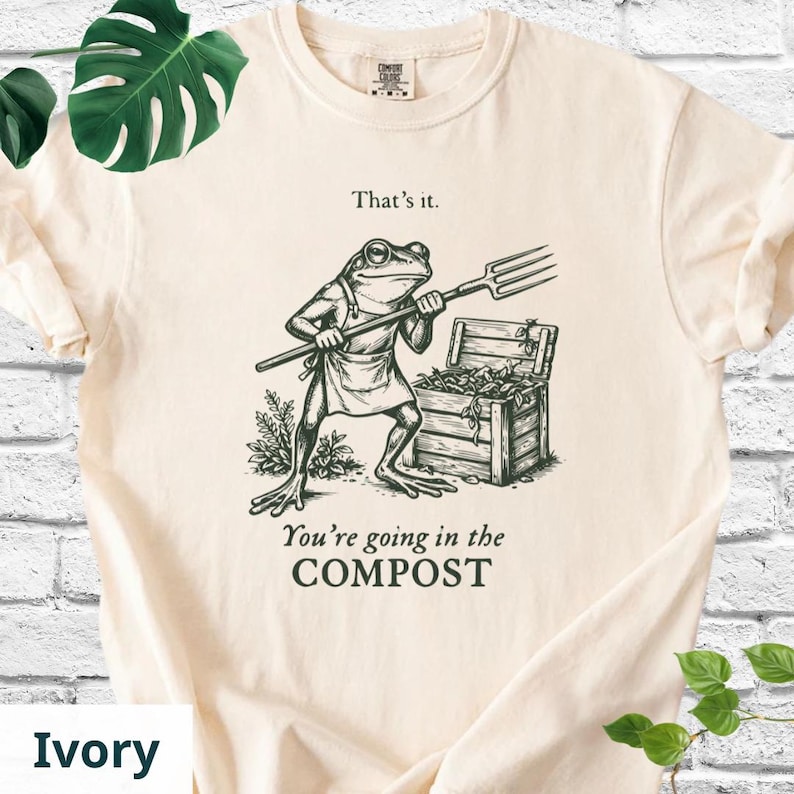 May include: Ivory-colored t-shirt featuring a humorous illustration of a frog holding a pitchfork, standing near a wooden compost bin. The text "That's it." and "You're going in the COMPOST" are printed above and below the frog graphic.