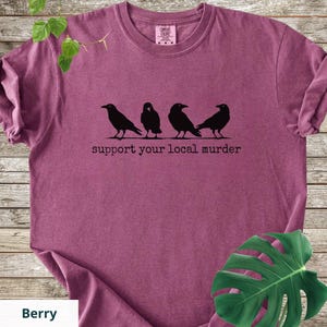 May include: A berry-colored short-sleeve t-shirt featuring a black graphic of four crows standing in a row. Below the birds, the text reads support your local murder in a typewriter-style font. The shirt has a crew neckline and a tag at the neck.