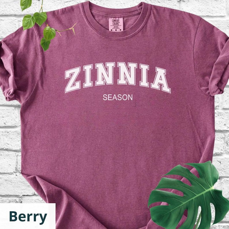 May include: Berry-coloured t-shirt with the word "ZINNIA" in white, arched lettering above the word "SEASON". The shirt is a Comfort Colors brand. A green leaf is in the lower right corner.