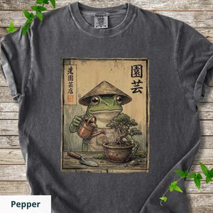 May include: Pepper-colored t-shirt featuring a whimsical illustration of a frog wearing a conical hat, watering a bonsai tree. The artwork includes Japanese text and a watering can. The shirt is made of a soft, comfortable material.