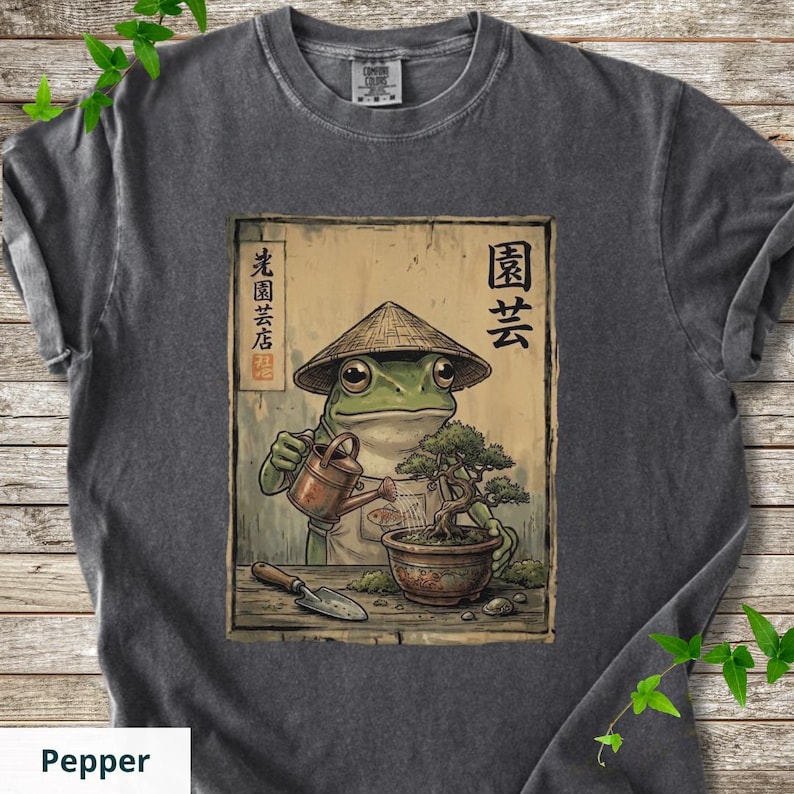 May include: Pepper-colored t-shirt featuring a whimsical illustration of a frog wearing a conical hat, watering a bonsai tree. The artwork includes Japanese text and a watering can. The shirt is made of a soft, comfortable material.