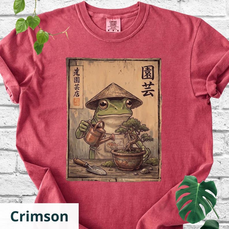 May include: Crimson-colored t-shirt featuring a frog wearing a conical hat, watering a bonsai tree. The frog illustration includes Japanese text and a small shovel. The shirt has a crew neck and short sleeves.