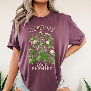 Peut inclure: T-shirt prune avec l'inscription "COMPOST YOUR ENEMIES" dans une police vintage. Le graphisme repr&eacute;sente trois squelettes parmi un feuillage vert, l'un tenant un arrosoir et l'autre une faux.