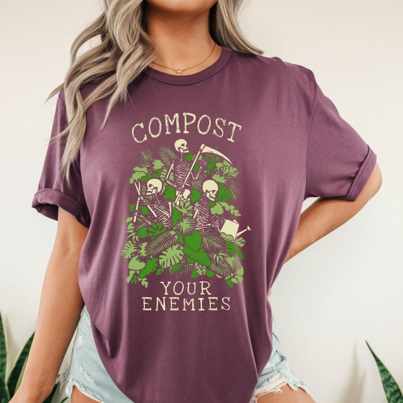 Peut inclure: T-shirt prune avec l'inscription "COMPOST YOUR ENEMIES" dans une police vintage. Le graphisme repr&eacute;sente trois squelettes parmi un feuillage vert, l'un tenant un arrosoir et l'autre une faux.