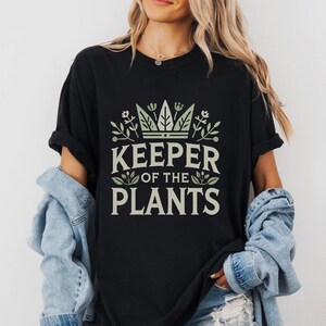 May include: Black t-shirt with the text "KEEPER OF THE PLANTS" in a light green font. The design includes a crown and botanical elements. The shirt has short sleeves and a relaxed fit.