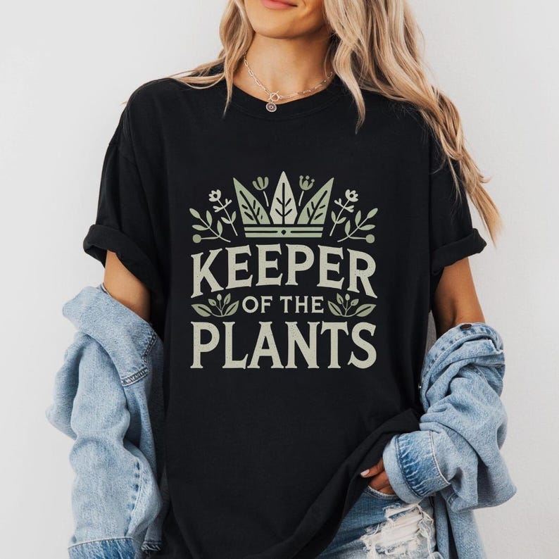 May include: Black t-shirt with the text "KEEPER OF THE PLANTS" in a light green font. The design includes a crown and botanical elements. The shirt has short sleeves and a relaxed fit.