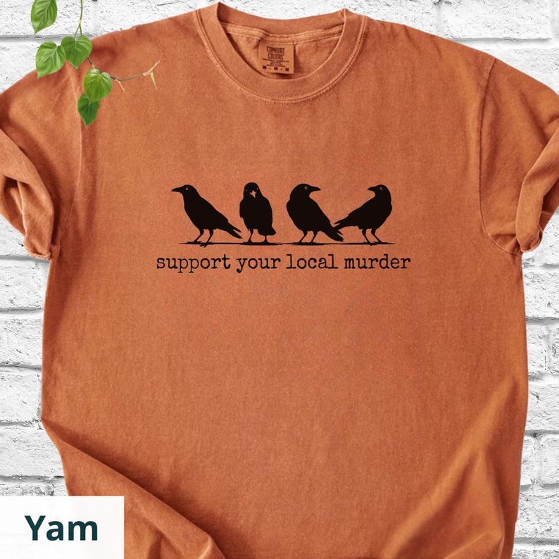 May include: A burnt orange short-sleeve t-shirt featuring a black graphic of four crows standing in a row. Below the birds, the text reads support your local murder in a typewriter-style font. The shirt has a crew neckline and a visible brand label.