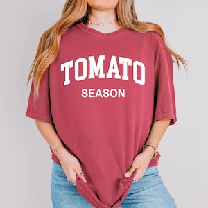 K&ouml;nnte beinhalten: Ein weinrotes T-Shirt mit dem wei&szlig;en Aufdruck "TOMATO SEASON" in gro&szlig;en Buchstaben. Das Shirt hat kurze &Auml;rmel und eine lockere Passform. Das Model tr&auml;gt eine blaue Jeans.