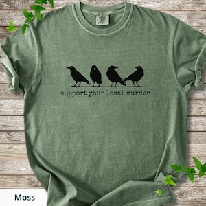 May include: A moss green short-sleeve t-shirt featuring a black graphic of four crows standing in a row. Below the birds, the text reads "support your local murder" in a typewriter-style font. The shirt has a crew neckline and a tag at the collar.