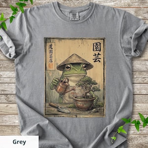 May include: Grey t-shirt featuring a frog in a conical hat watering a bonsai tree. The frog is depicted in a vintage style, with Japanese text and a gardening theme. The word "Grey" is at the bottom of the shirt.