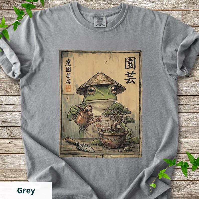 May include: Grey t-shirt featuring a frog in a conical hat watering a bonsai tree. The frog is depicted in a vintage style, with Japanese text and a gardening theme. The word "Grey" is at the bottom of the shirt.