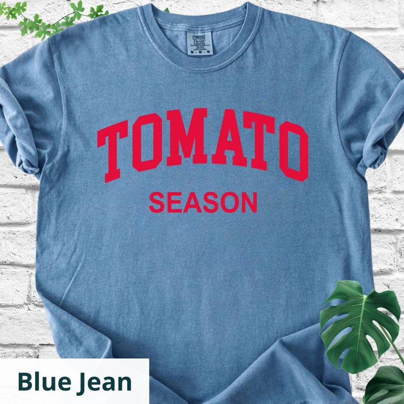 K&ouml;nnte beinhalten: Blaues Jeans-T-Shirt mit den Worten "TOMATO SEASON" in roten Blockbuchstaben. Das Shirt hat einen Rundhalsausschnitt und kurze &Auml;rmel. Das Shirt ist aus einem weichen, bequemen Material.