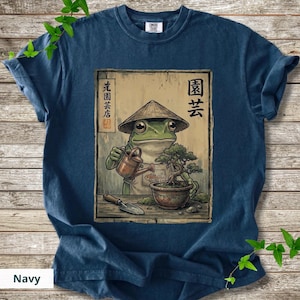 May include: Navy blue t-shirt featuring a frog wearing a conical hat, watering a bonsai tree. The illustration includes Japanese text and a small shovel. The shirt is a comfortable, casual wear.