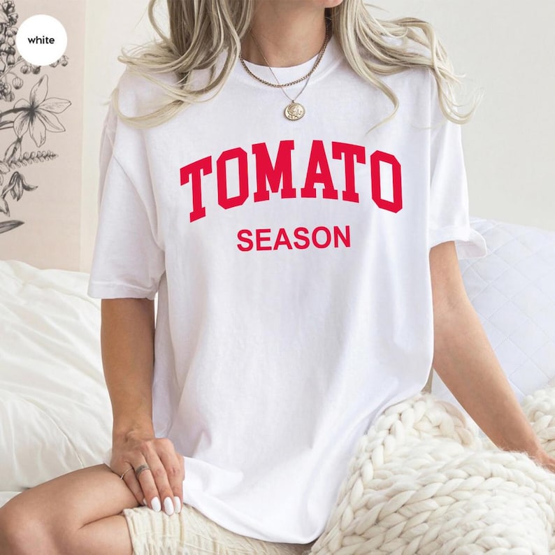 K&ouml;nnte beinhalten: Ein wei&szlig;es T-Shirt mit dem roten Aufdruck "TOMATO SEASON". Das Shirt hat einen l&auml;ssigen Stil mit kurzen &Auml;rmeln und einer lockeren Passform. Der Text ist mittig auf der Vorderseite des Shirts platziert.