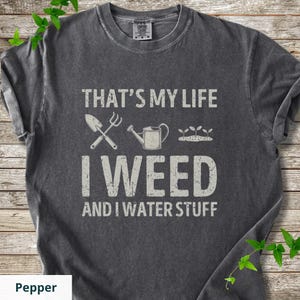 May include: A charcoal gray short-sleeve t-shirt with a distressed white graphic print. The text reads THAT'S MY LIFE I WEED AND I WATER STUFF, accompanied by illustrations of a hand trowel, a watering can, and three small sprouting plants.