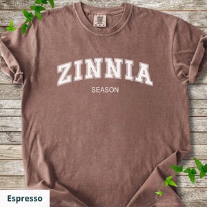 May include: Espresso-coloured t-shirt featuring the word "ZINNIA" in white, arched lettering above the word "SEASON". The shirt has a crew neck and short sleeves. The shirt is displayed on a wooden surface.