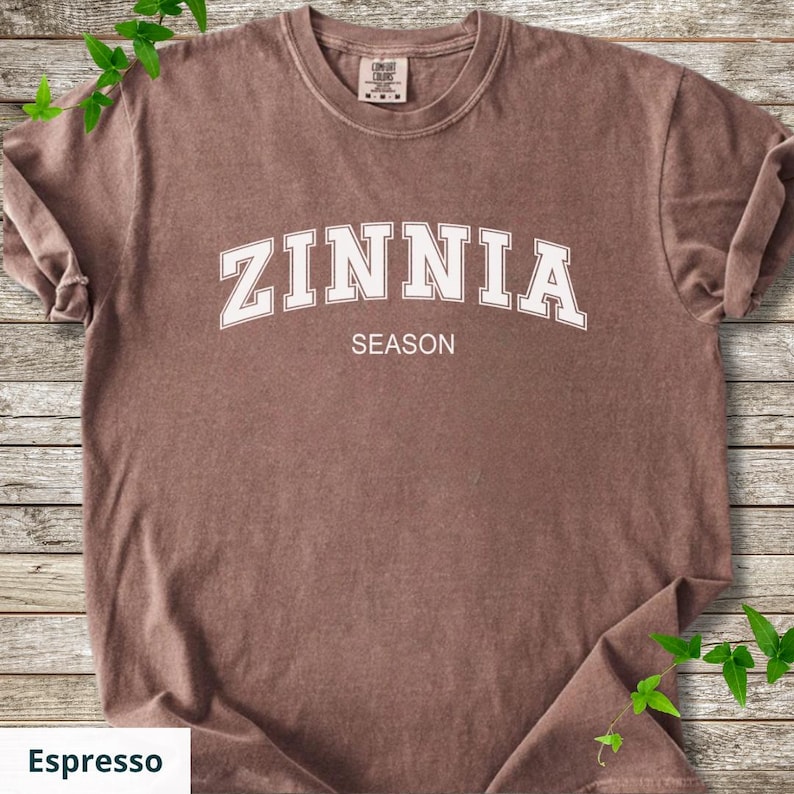 May include: Espresso-coloured t-shirt featuring the word "ZINNIA" in white, arched lettering above the word "SEASON". The shirt has a crew neck and short sleeves. The shirt is displayed on a wooden surface.