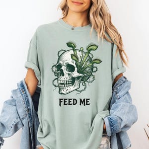 May include: Sage green t-shirt featuring a detailed skull graphic with Venus flytraps and thorny vines. The text "FEED ME" is printed in bold, black letters below the skull. The shirt has a relaxed fit.