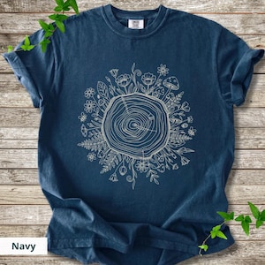 May include: Navy blue t-shirt featuring a cream-colored graphic of a tree trunk cross-section surrounded by flowers, mushrooms, and foliage. The shirt has short sleeves and a crew neck. The word "Navy" is printed at the bottom.