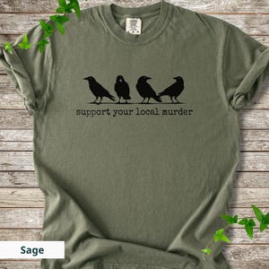 May include: A sage green short-sleeve t-shirt featuring a black graphic of four crows standing in a row. Below the birds, the text reads "support your local murder" in a typewriter-style font. The shirt has a crew neckline and a tag at the collar.