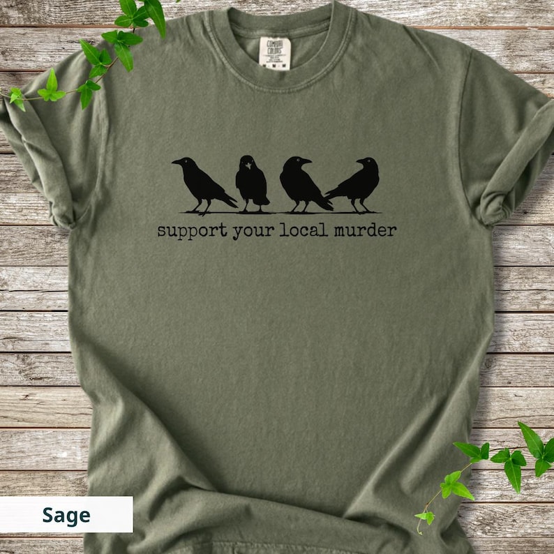 May include: A sage green short-sleeve t-shirt featuring a black graphic of four crows standing in a row. Below the birds, the text reads "support your local murder" in a typewriter-style font. The shirt has a crew neckline and a tag at the collar.