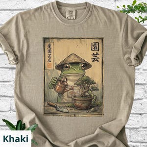 May include: Khaki-colored t-shirt featuring a frog wearing a conical hat, watering a bonsai tree. The frog is depicted in a Japanese art style. The shirt has the text "Comfort Colors" on the tag.
