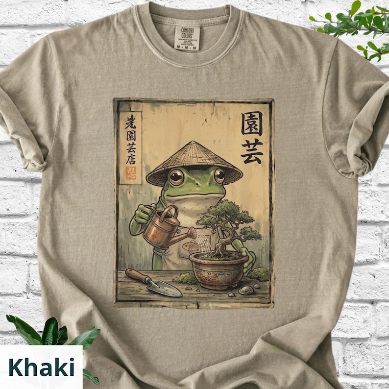 May include: Khaki-colored t-shirt featuring a frog wearing a conical hat, watering a bonsai tree. The frog is depicted in a Japanese art style. The shirt has the text "Comfort Colors" on the tag.