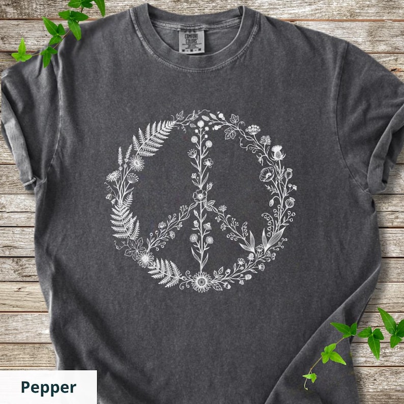 May include: A charcoal gray short-sleeve t-shirt featuring a white graphic print of a peace sign composed of various botanical elements, including ferns, wildflowers, and leafy stems. The shirt has a crew neckline and a vintage-style wash.