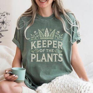 May include: Sage green t-shirt with the text "KEEPER OF THE PLANTS" in a light green font. The text is surrounded by a crown and floral design. The shirt has rolled-up sleeves.