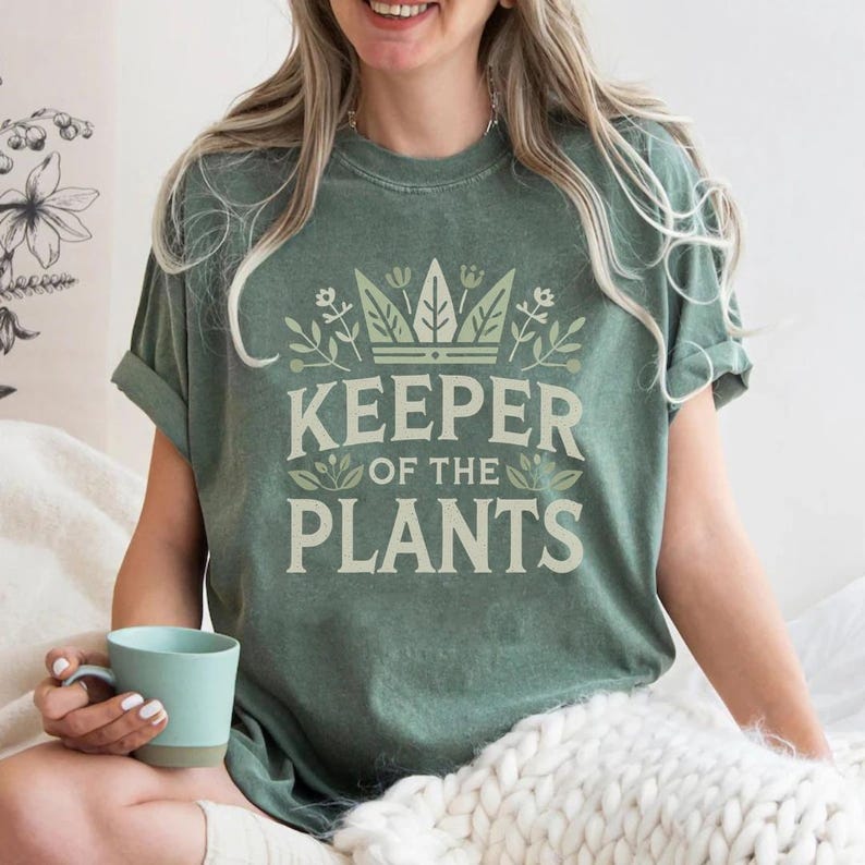 May include: Sage green t-shirt with the text "KEEPER OF THE PLANTS" in a light green font. The text is surrounded by a crown and floral design. The shirt has rolled-up sleeves.