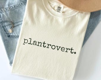 May include: An ivory t-shirt with the word "plantrovert" printed in dark green. The shirt is folded and rests on light blue jeans. A tan hat and a silver necklace are also visible.