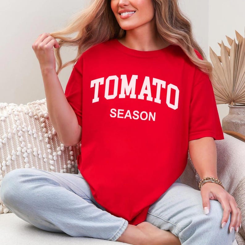K&ouml;nnte beinhalten: Ein leuchtend rotes T-Shirt mit dem Aufdruck "TOMATO SEASON" in gro&szlig;en, wei&szlig;en Buchstaben. Das Shirt hat eine einfarbige Gestaltung und scheint aus einem weichen Material zu bestehen. Es ist ein l&auml;ssiges Kleidungsst&uuml;ck.