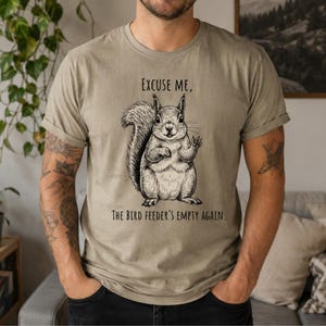 May include: A light olive green t-shirt featuring a detailed black and white illustration of a squirrel. The squirrel is waving and the text reads "Excuse me, the bird feeder's empty again."