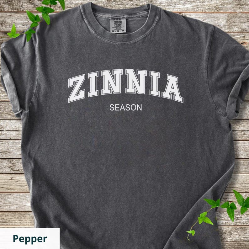 May include: A charcoal grey short-sleeve t-shirt with a crew neckline. The chest features the white text ZINNIA in an arched collegiate font, with the word SEASON printed in a smaller, straight white sans-serif font below it.