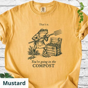 May include: Mustard-colored t-shirt featuring a frog character holding a pitchfork, standing near a wooden chest. The text "That's it." and "You're going in the COMPOST" are printed on the shirt. The shirt is made of a soft material.