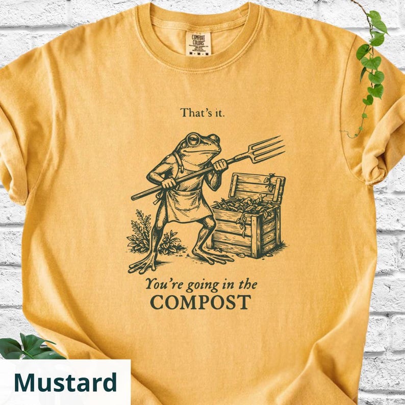 May include: Mustard-colored t-shirt featuring a frog character holding a pitchfork, standing near a wooden chest. The text "That's it." and "You're going in the COMPOST" are printed on the shirt. The shirt is made of a soft material.