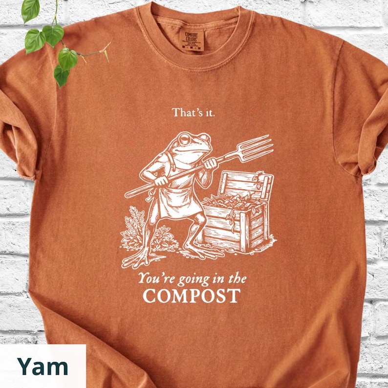 May include: A burnt orange t-shirt featuring a white illustration of a frog holding a pitchfork, standing near a wooden compost bin. The text "That's it." and "You're going in the COMPOST" are printed on the shirt.
