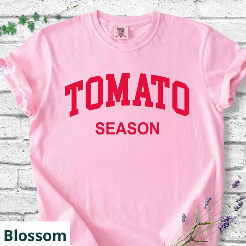 K&ouml;nnte beinhalten: Hellrosa T-Shirt mit dem roten Aufdruck "TOMATO SEASON". Das T-Shirt hat hochgekrempelte &Auml;rmel und besteht aus einem weichen Stoff. Die Farbe des T-Shirts ist Blossom.