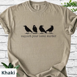 May include: A khaki-colored crew neck t-shirt featuring a black graphic of four crows standing in a row. Below the birds, the text reads "support your local murder" in a typewriter-style font. The shirt has a ribbed collar and short sleeves.