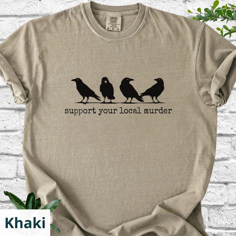 May include: A khaki-colored crew neck t-shirt featuring a black graphic of four crows standing in a row. Below the birds, the text reads "support your local murder" in a typewriter-style font. The shirt has a ribbed collar and short sleeves.